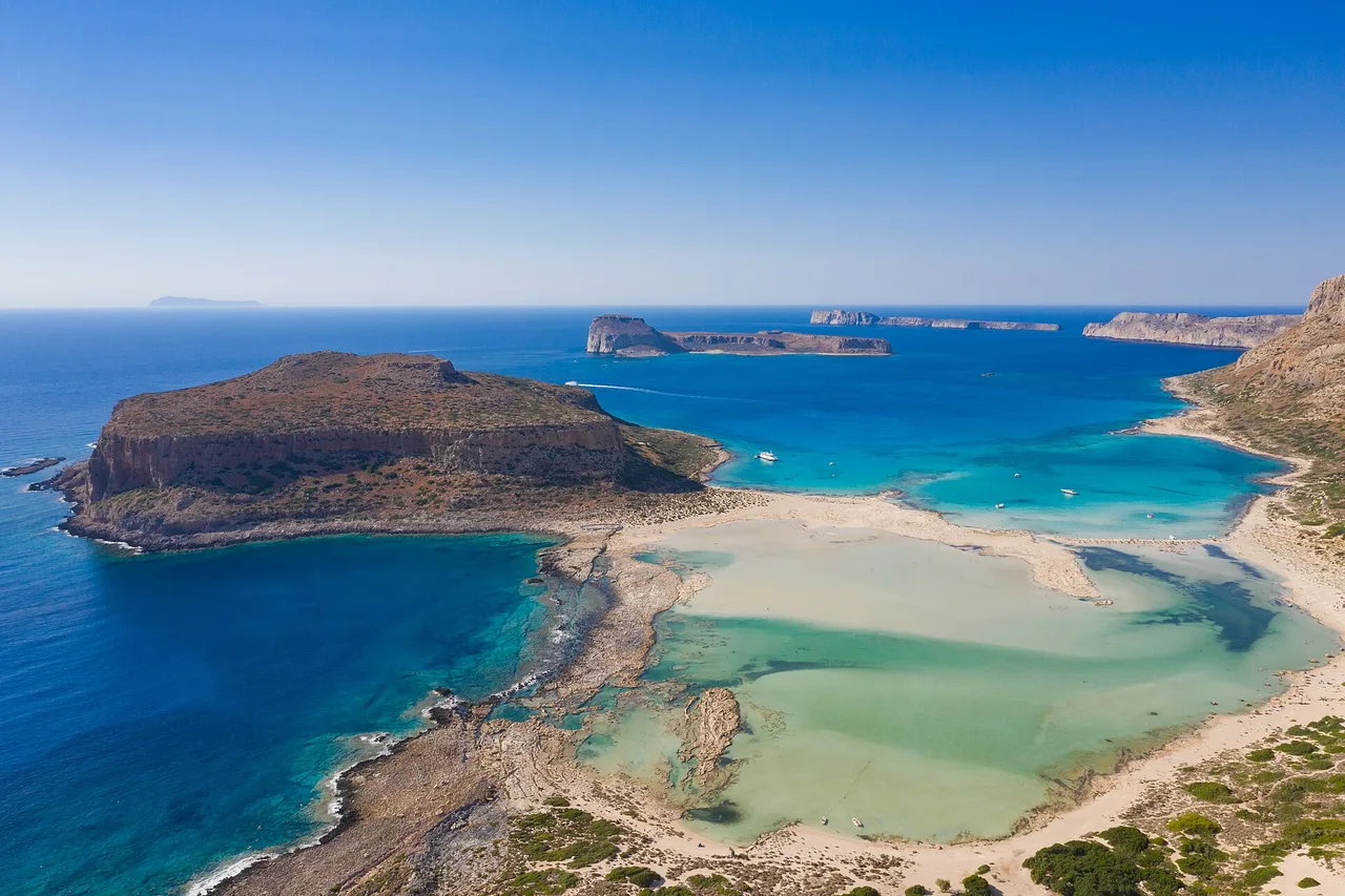Vista aérea de la laguna de Balos: bancos de arena clara, agua turquesa muy somera, a la izquierda la península rocosa de Gramvusa y, mar adentro, el islote fortificado.