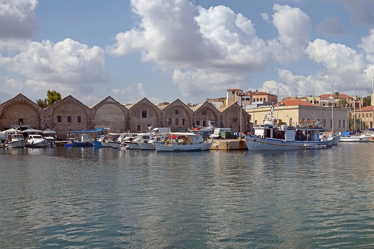El puerto veneciano de La Canea: barcas de pesca y veleros amarrados en el muelle, fachadas ocres y rojas de las casas del paseo marítimo, bajo un cielo de grandes nubes blancas.