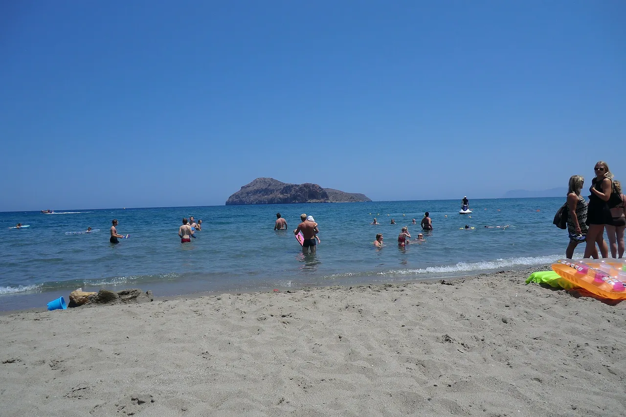 Plage de sable d’Agia Marina, à l’ouest de La Canée : des baigneurs dans une eau claire et peu profonde, et au large le petit îlot rocheux d’Agii Theodori.