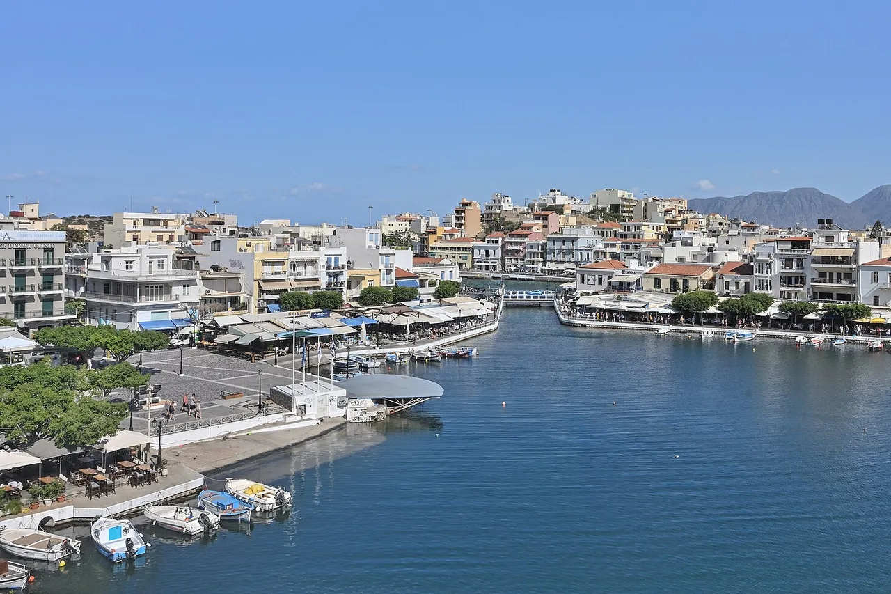 Le lac Voulismeni à Agios Nikolaos vu d’en haut : un port rond d’un bleu profond avec de petits bateaux, bordé de cafés et de maisons blanches, montagnes au fond.