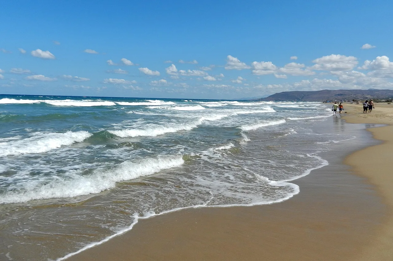 La longue et large plage de sable de Georgioupoli, sur la côte nord de la Crète, avec des vagues qui s’étalent, quelques promeneurs et des montagnes à l’horizon.