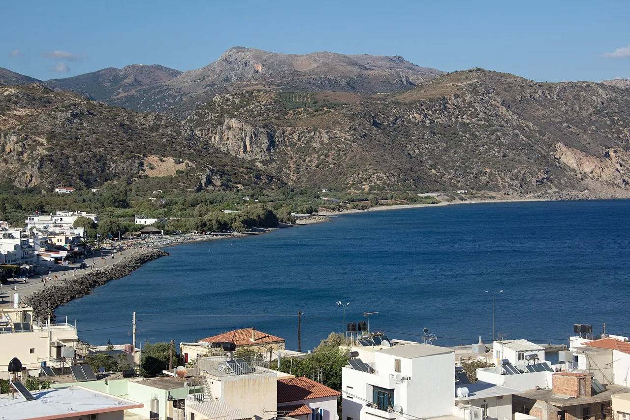Paleochora, sur la côte sud-ouest de la Crète, vue d’en haut : maisons blanches au premier plan, une jetée, la baie d’un bleu profond et des montagnes nues et abruptes derrière.