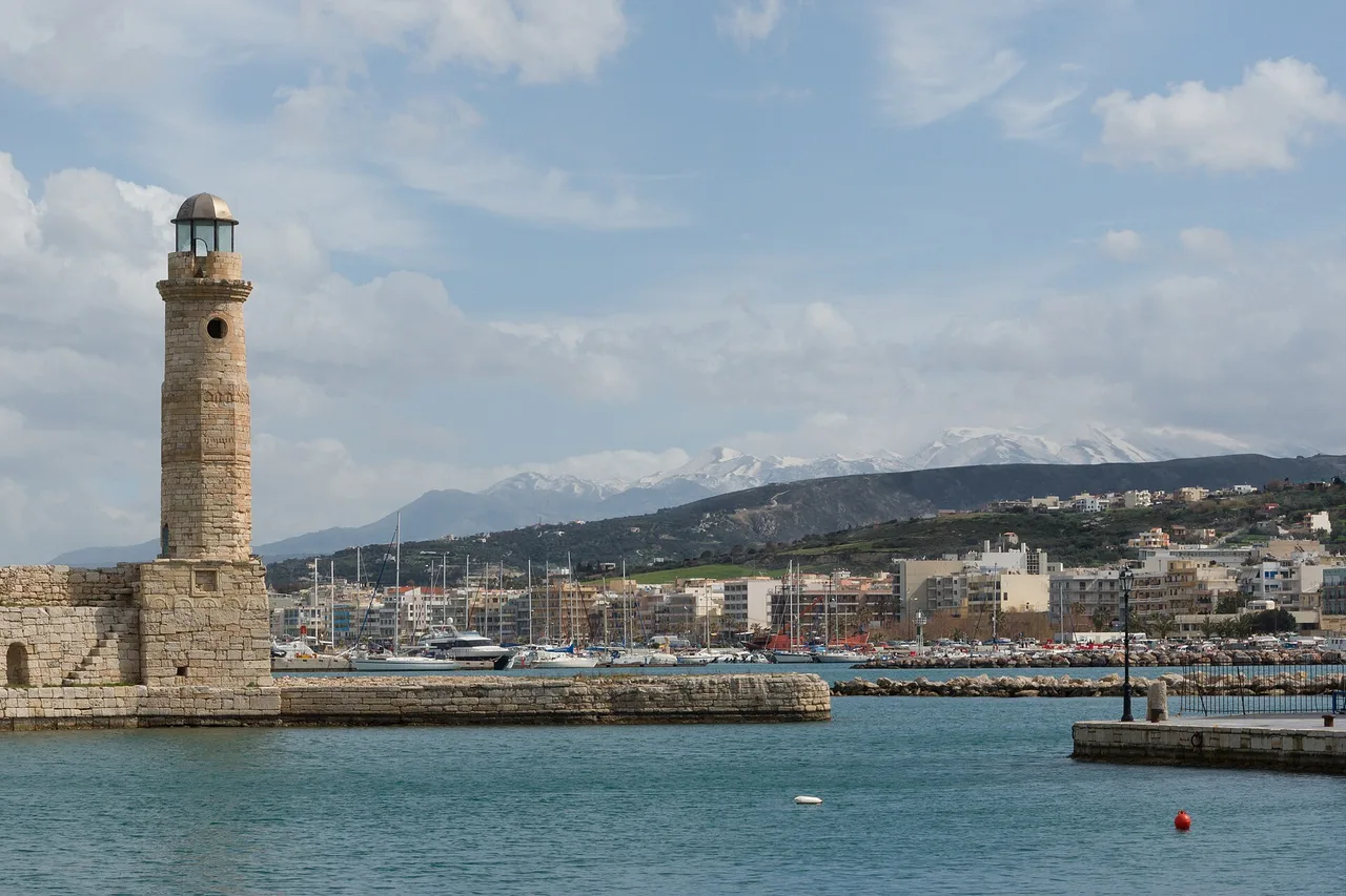 Le port vénitien de Réthymnon, en Crète : à gauche le phare de pierre sur la jetée, derrière les bateaux et les façades du front de mer, et à l’horizon les Montagnes Blanches enneigées.