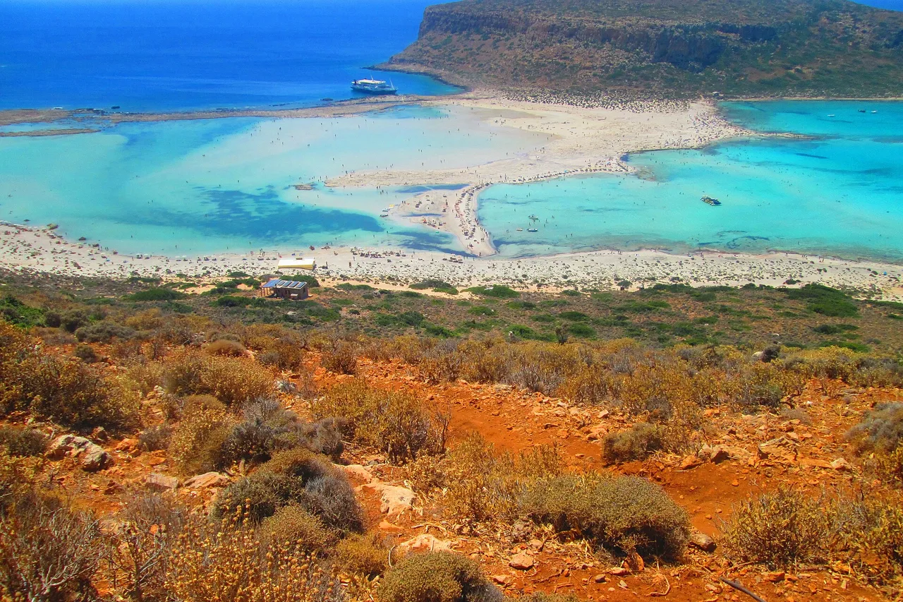 Vue aérienne de la lagune de Balos, en Crète : au premier plan une colline pelée d’un rouge rouille couverte de broussailles basses, derrière elle la lagune turquoise, sa langue de sable clair et l’îlot rocheux du large.