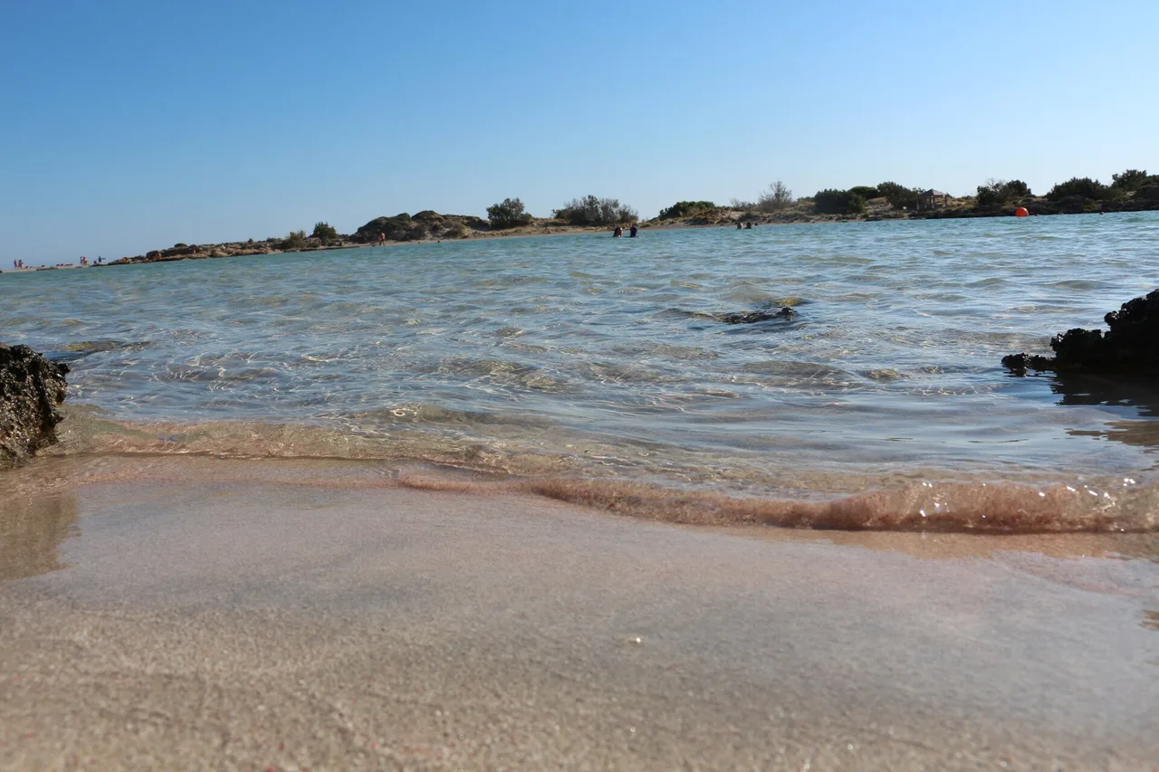 Gros plan de la ligne d’eau à Elafonissi : une bande de sable nettement rosée au bord de la vague qui se retire, tandis que le sable voisin reste clair et beige.