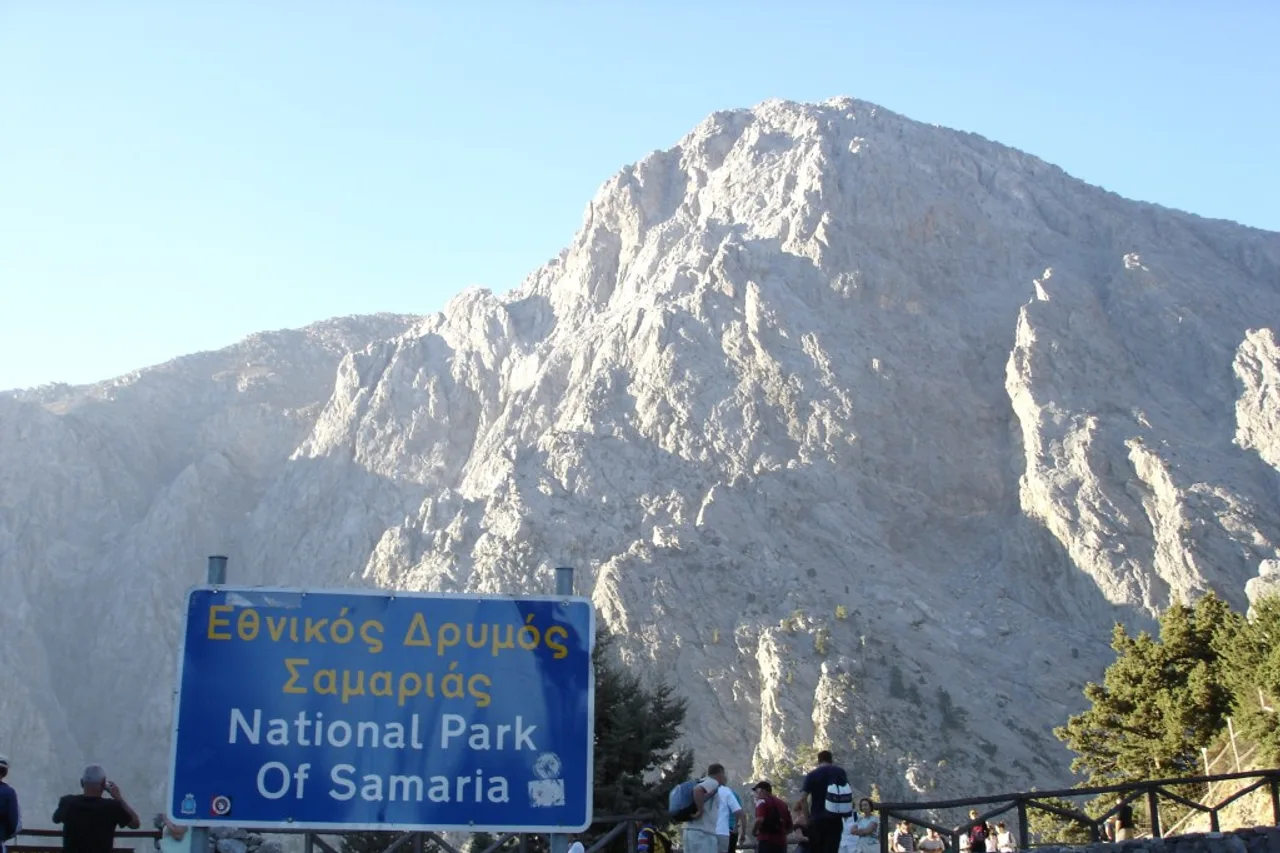Le panneau bleu « Parc national de Samaria » en grec et en anglais à l’entrée de Xyloskalo, avec derrière des randonneurs le long d’une rambarde et un sommet rocheux clair et découpé.