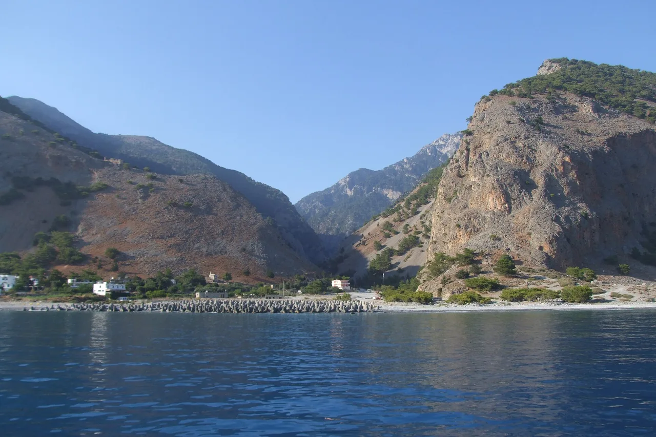 Agia Rouméli vue de la mer : quelques maisons blanches au bord d’une plage de galets, et derrière le débouché de la gorge entre de hautes montagnes nues.