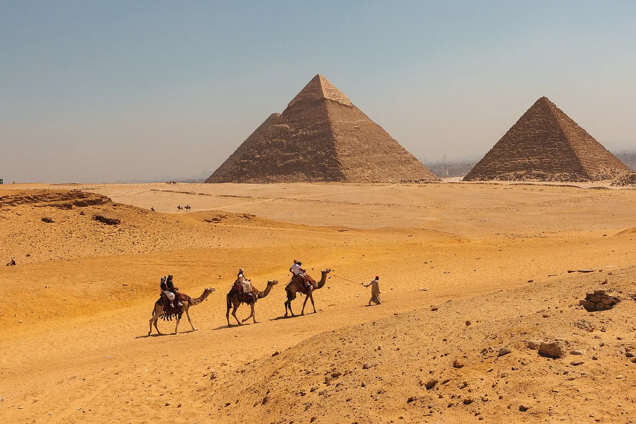 Una piccola carovana di cammelli attraversa la sabbia, con due delle piramidi di Giza sullo sfondo sotto un cielo chiaro.
