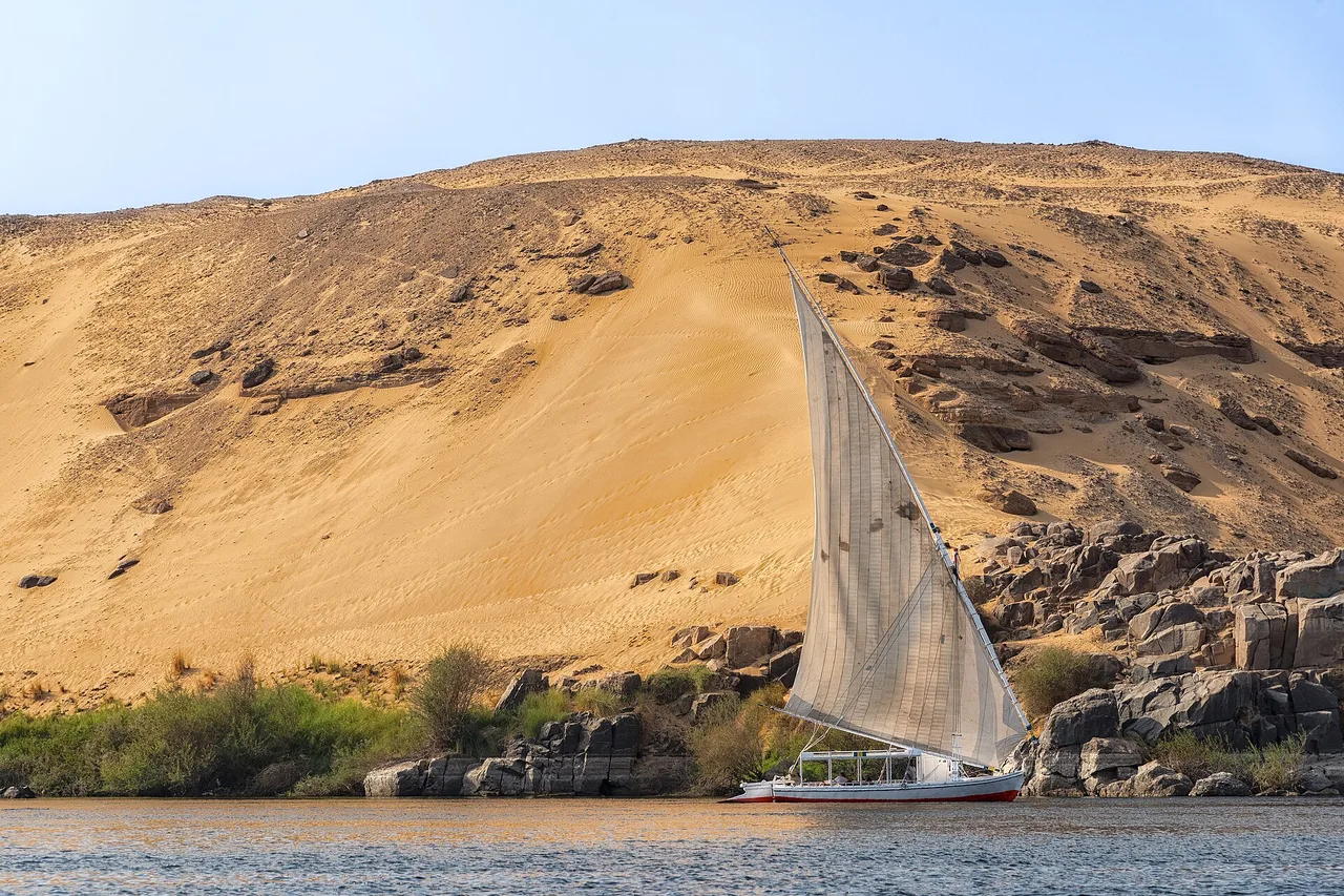 Une felouque à voile blanche glisse sur le Nil devant une haute dune de sable et des rochers sombres.