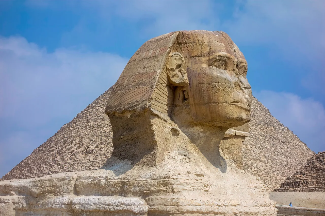 La tête du Sphinx de Gizeh en gros plan, une pyramide en arrière-plan sous un ciel bleu.