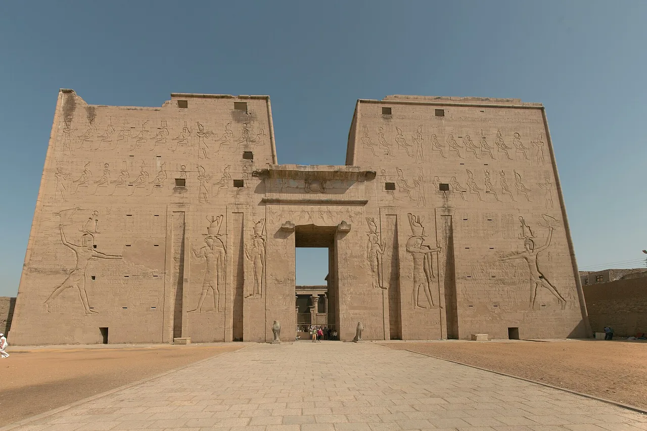 Le haut pylône d'entrée du temple d'Horus à Edfou, couvert de reliefs, devant un parvis dallé.