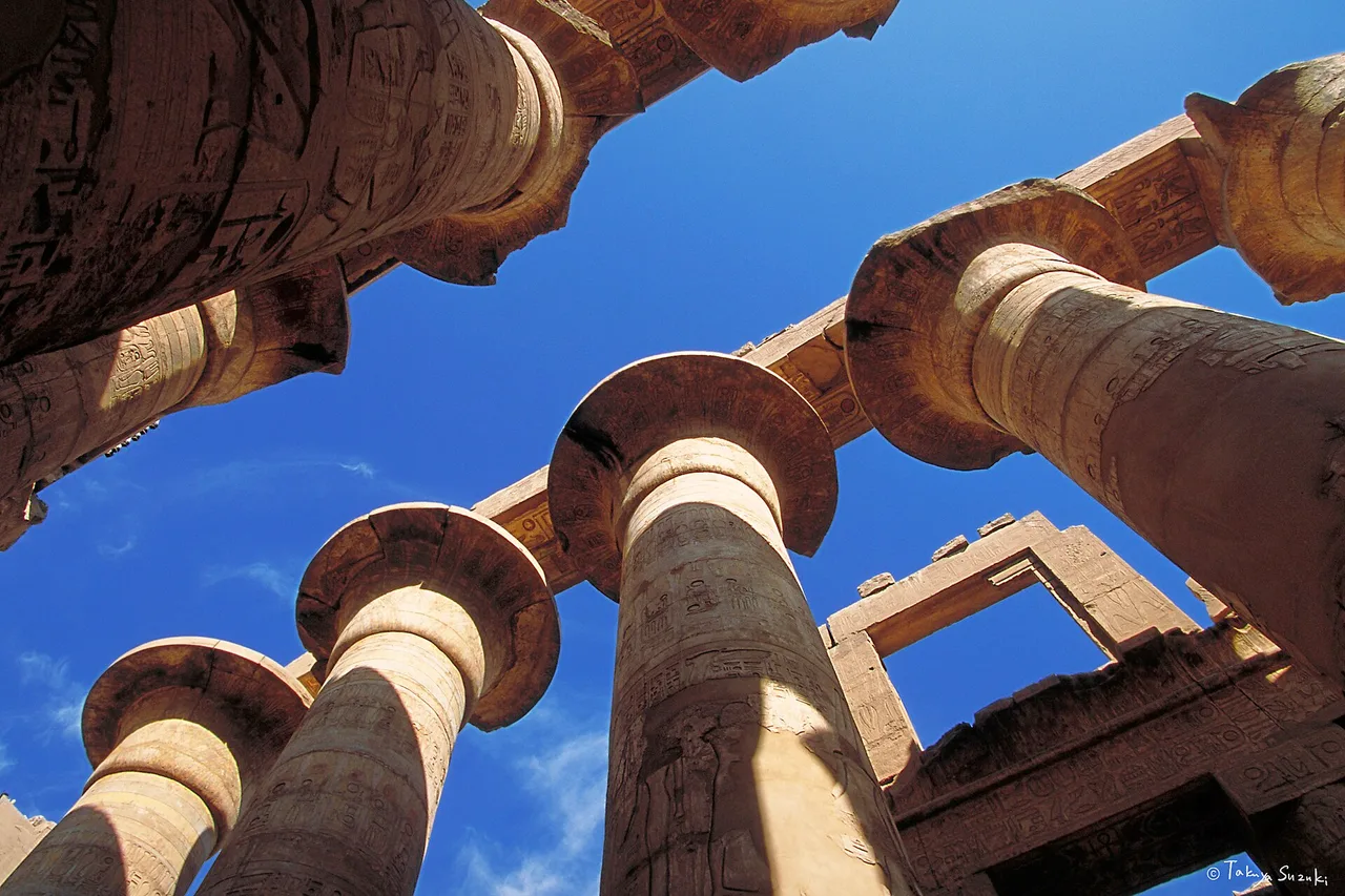 Vue en contre-plongée des immenses colonnes papyriformes de la grande salle hypostyle de Karnak sur fond de ciel bleu.