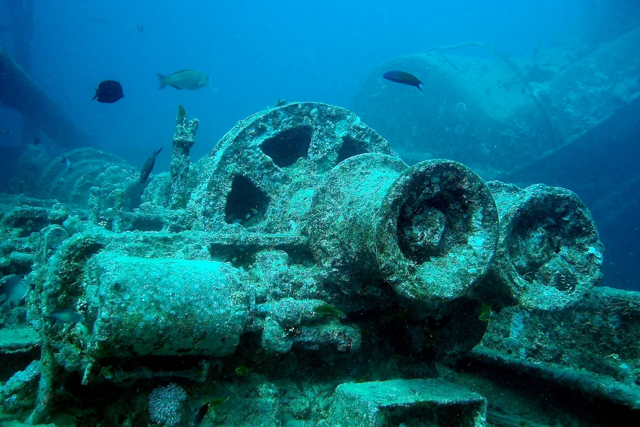 Unterwasseraufnahme von Lokomotivteilen auf dem Deck des Wracks SS Thistlegorm im Roten Meer, umgeben von Fischen.