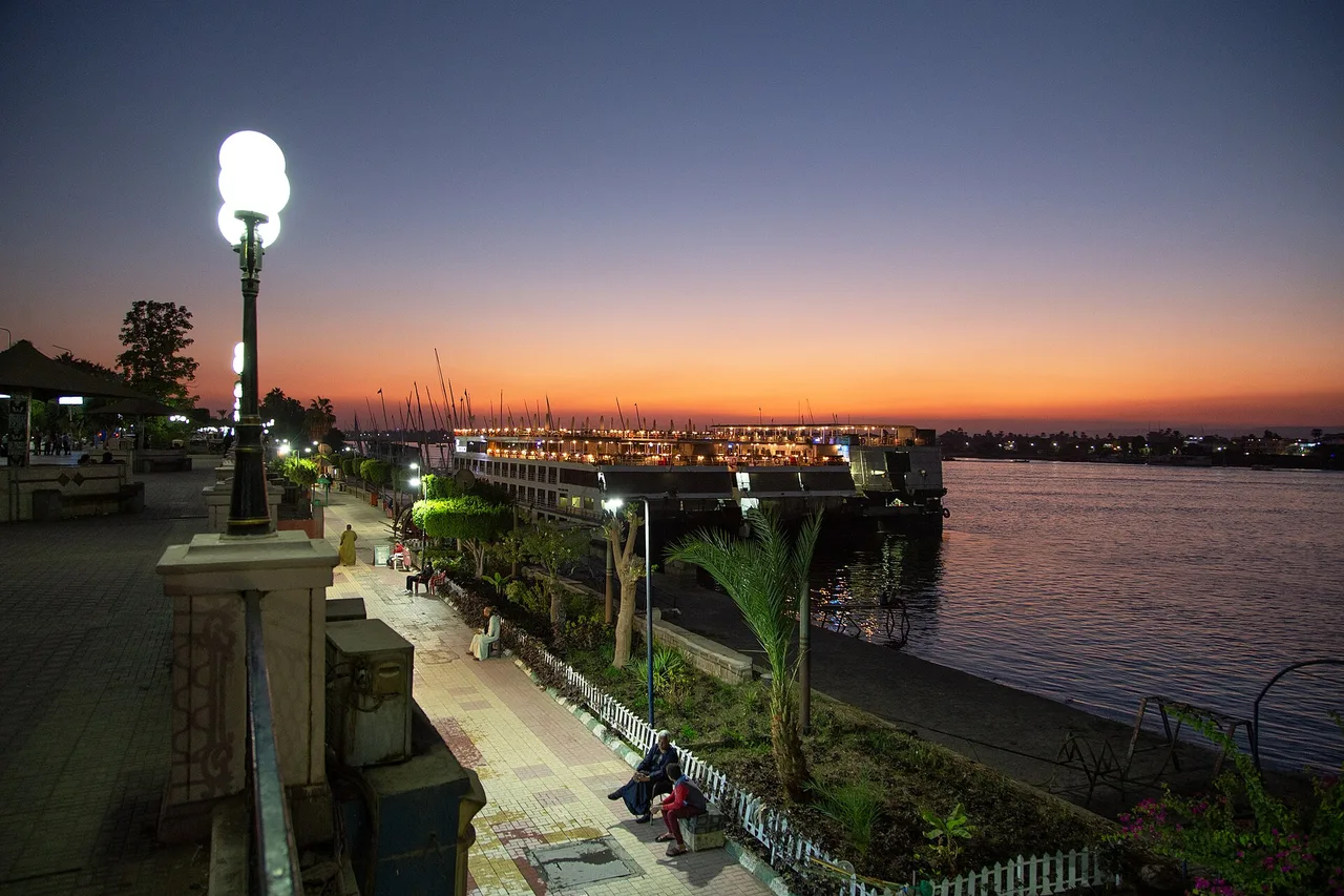 The illuminated Luxor corniche on the Nile at dusk, with street lamps and boats moored along the quay.