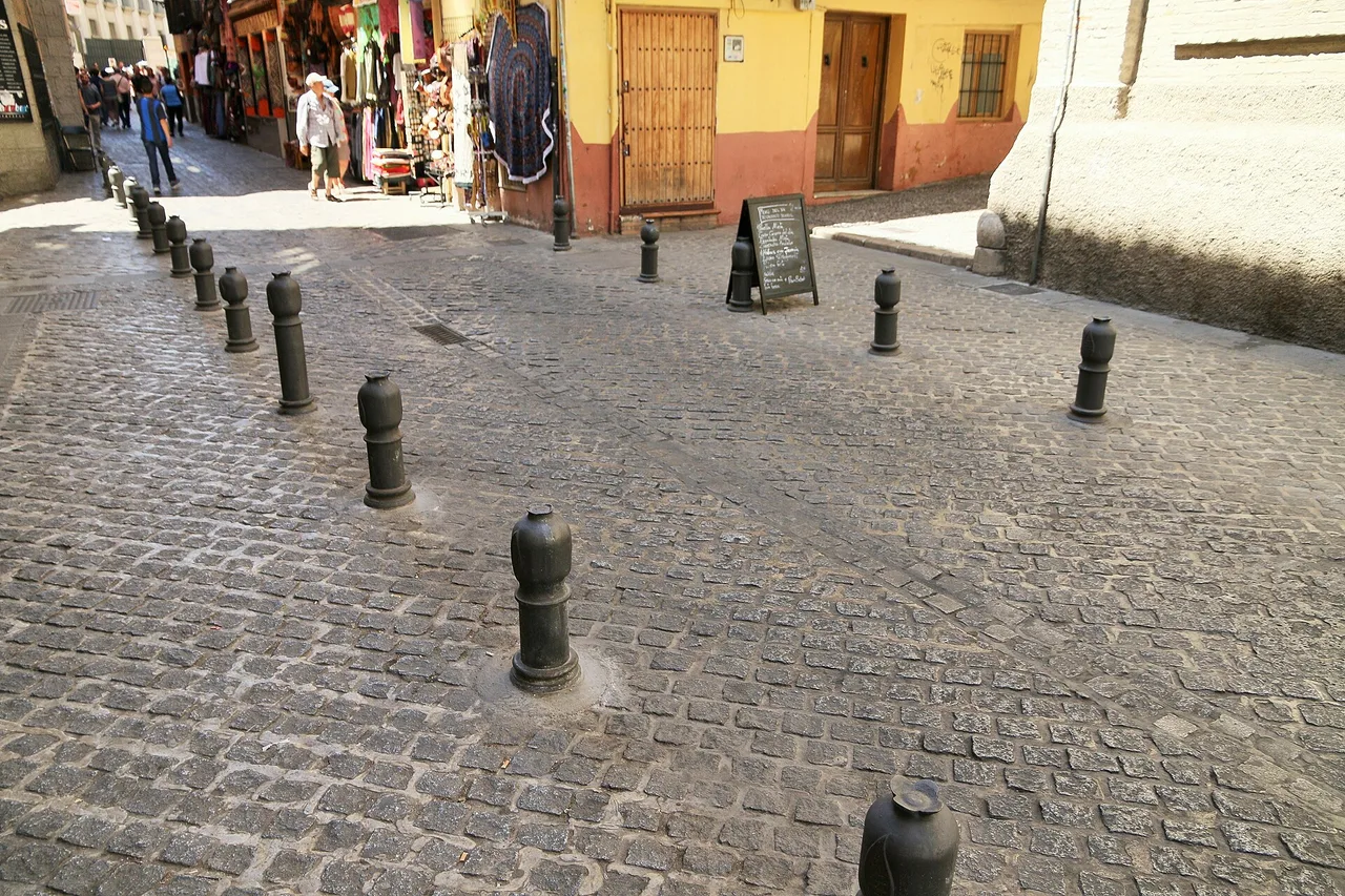 Schwarze Poller auf einer Kopfsteinpflasterstraße im Viertel Albaicín in Granada, im Hintergrund Geschäfte, eine Kreidetafel und Passanten
