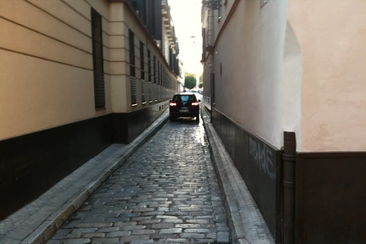 Ein dunkles Auto fährt mit eingeschalteten Rücklichtern durch eine sehr enge Kopfsteinpflastergasse in Sevilla, links und rechts hohe Hauswände ohne Platz zum Ausweichen