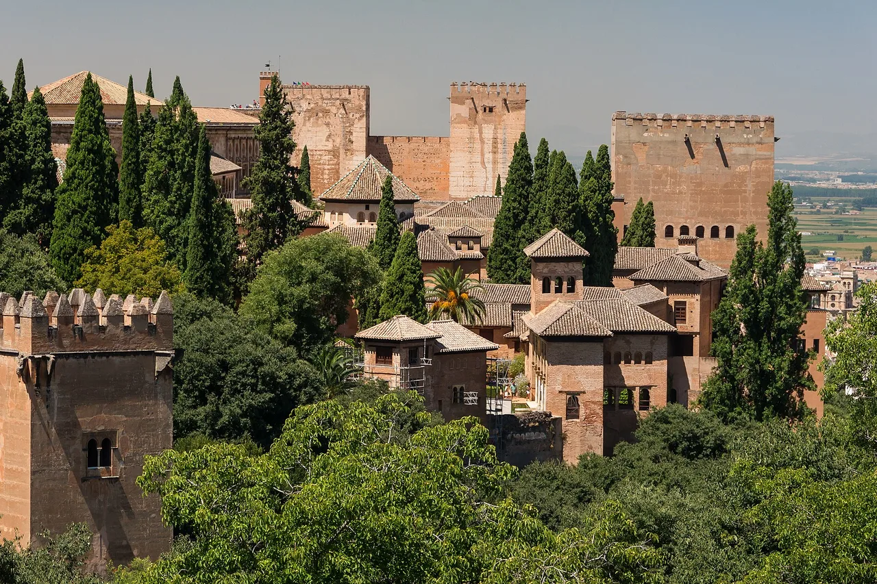 Des toits de tuiles et des tours crénelées de l’Alhambra émergent d’une végétation dense de cyprès, vus depuis les jardins du Generalife à Grenade