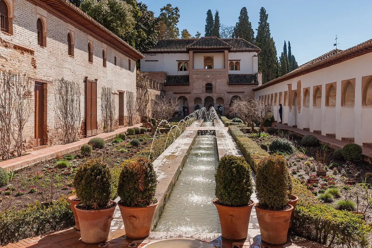 Un étroit canal d’eau à petits jets traverse le jardin du Generalife en direction d’un bâtiment du palais, avec des massifs et des pots en terre cuite sur les côtés