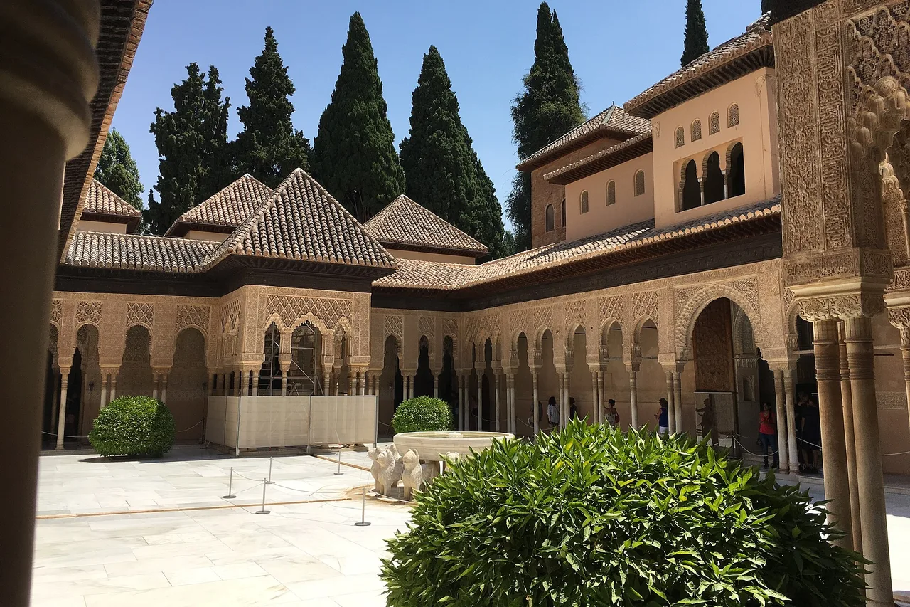 La cour des Lions de l’Alhambra en plein jour : une galerie de colonnes aux arcs ajourés, la fontaine au centre, des cyprès et un ciel bleu en arrière-plan