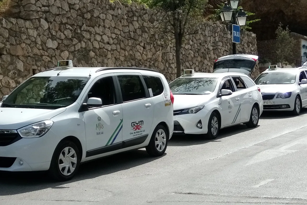 Taxis blancs garés le long d’un mur de pierre sur la Costa del Sol, un monospace Dacia au premier plan avec une inscription sur la portière