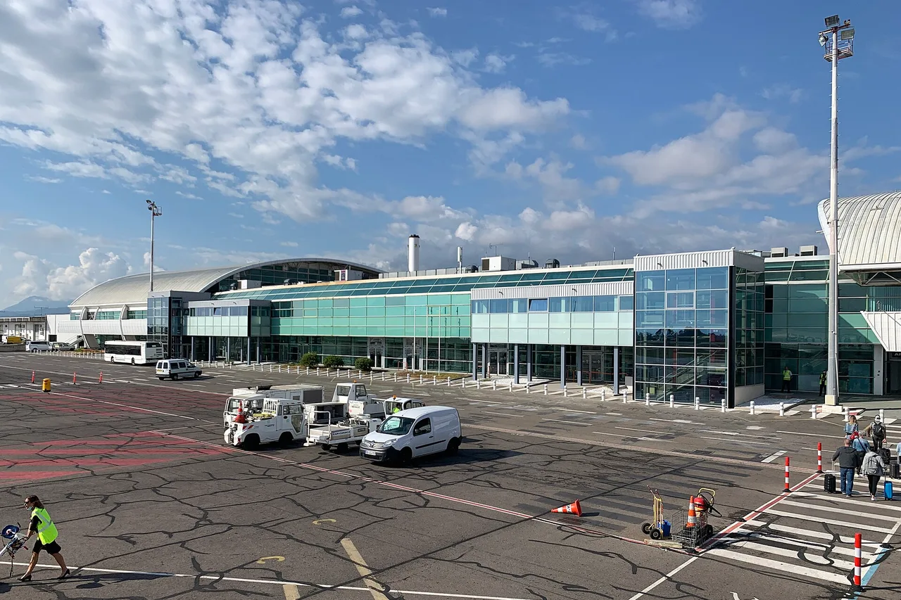 Il lungo terminal a vetri dell’aeroporto di Bastia-Poretta, con carrelli portabagagli e un furgone bianco sul piazzale, viaggiatori sulla destra