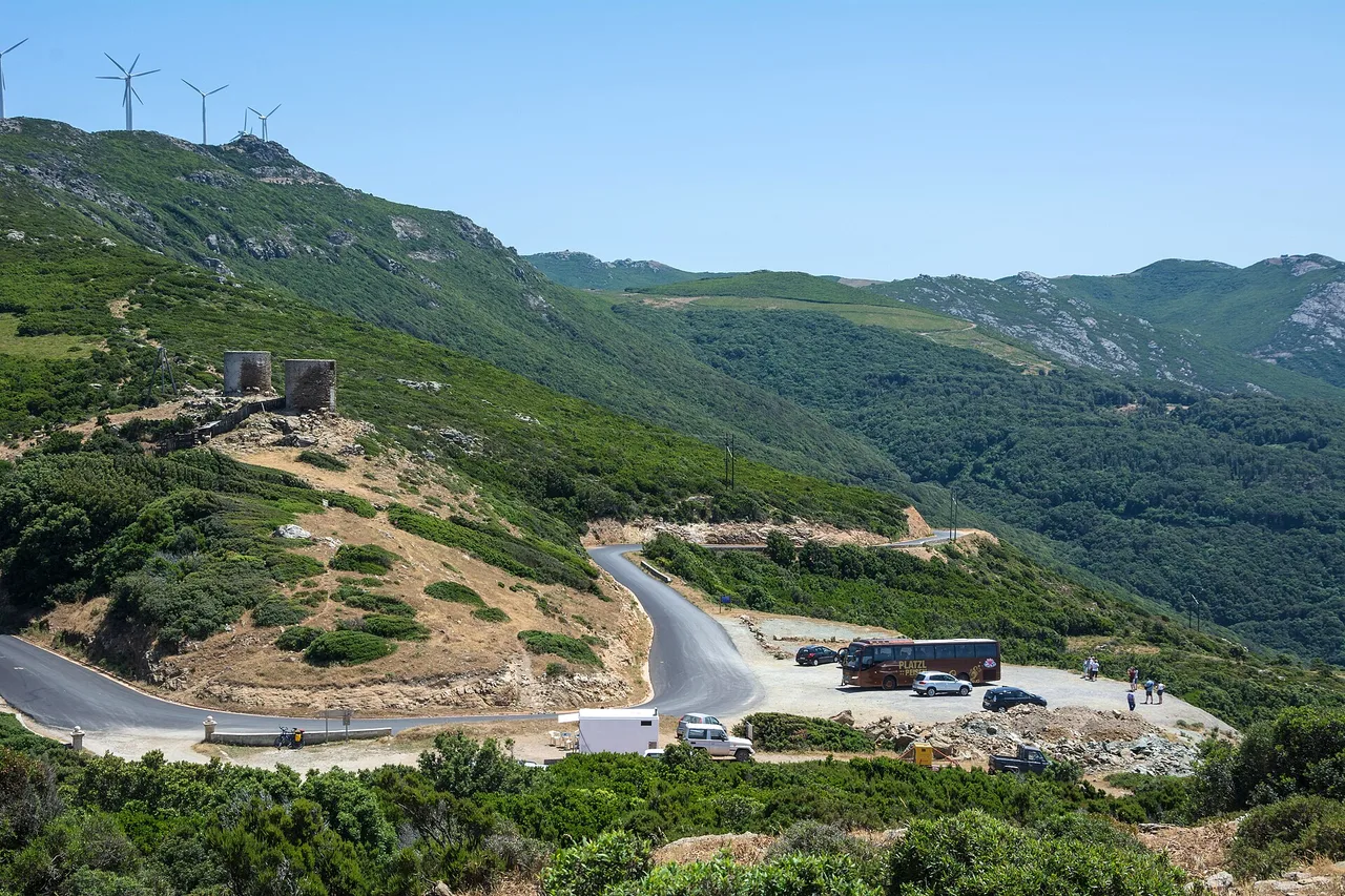 Una strada sale a tornanti su un versante verde del Capo Corso, sul crinale girano le pale eoliche, accanto a una torre in rovina e a un parcheggio con veicoli