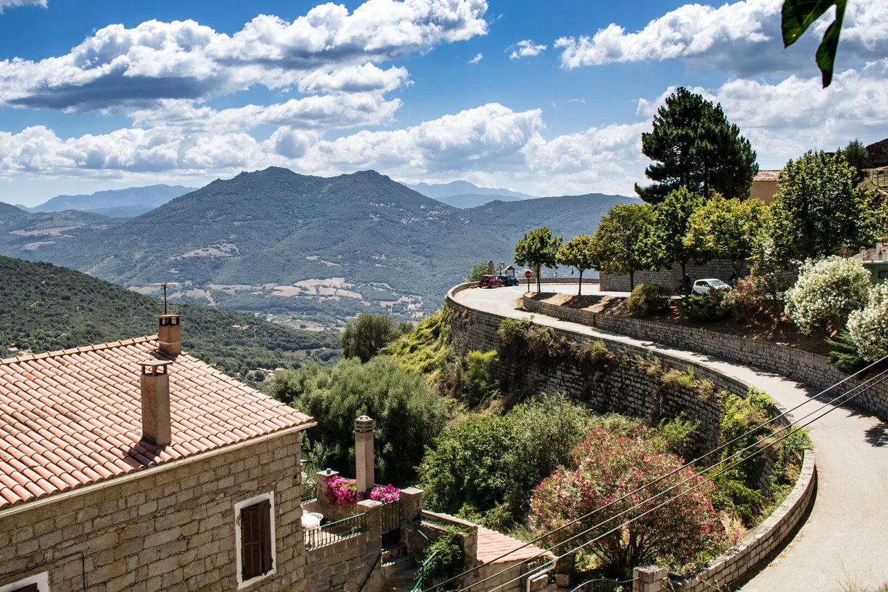 Una strada stretta con un basso muretto di pietra si curva su un versante sopra un paese corso, montagne sullo sfondo e un tetto di tegole in primo piano