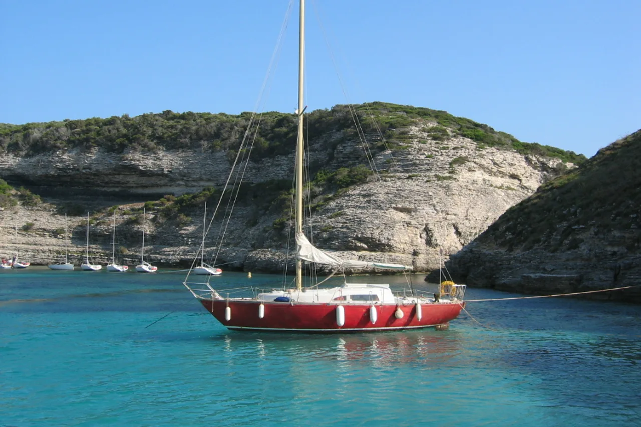 Una barca a vela dallo scafo rosso ancorata in acque turchesi davanti alle bianche falesie calcaree di Bonifacio.