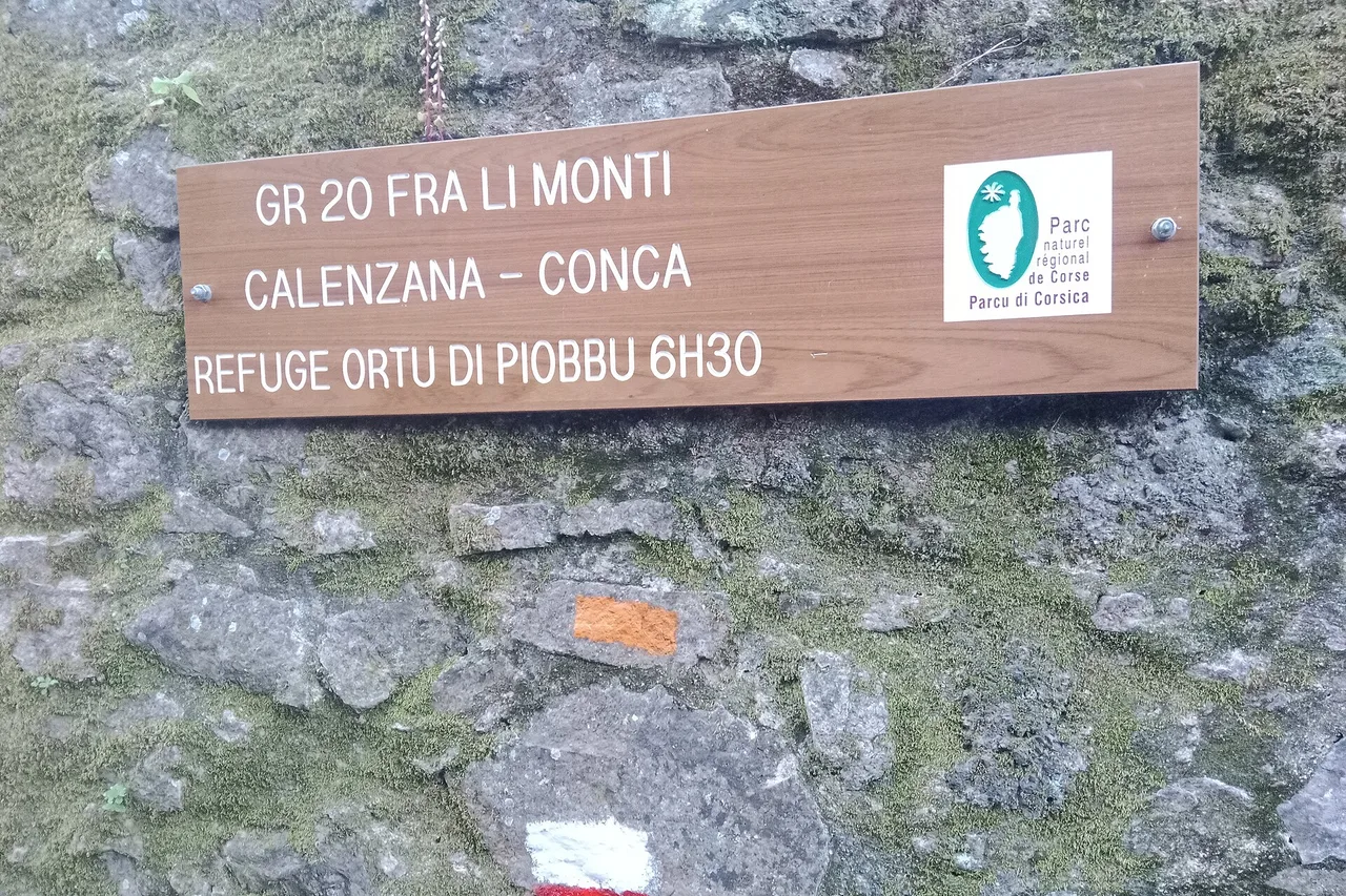 Un cartello di legno alla partenza del GR20 a Calenzana indica 6 h 30 fino al rifugio di Ortu di u Piobbu; sotto, un segnavia arancione su un muro di pietra.