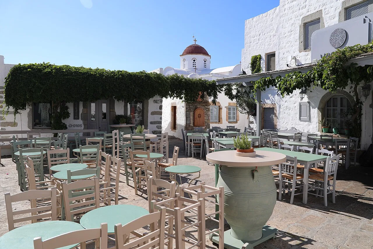 Une placette blanche de la chora de Patmos, chaises et tables de taverne rangées sous une treille