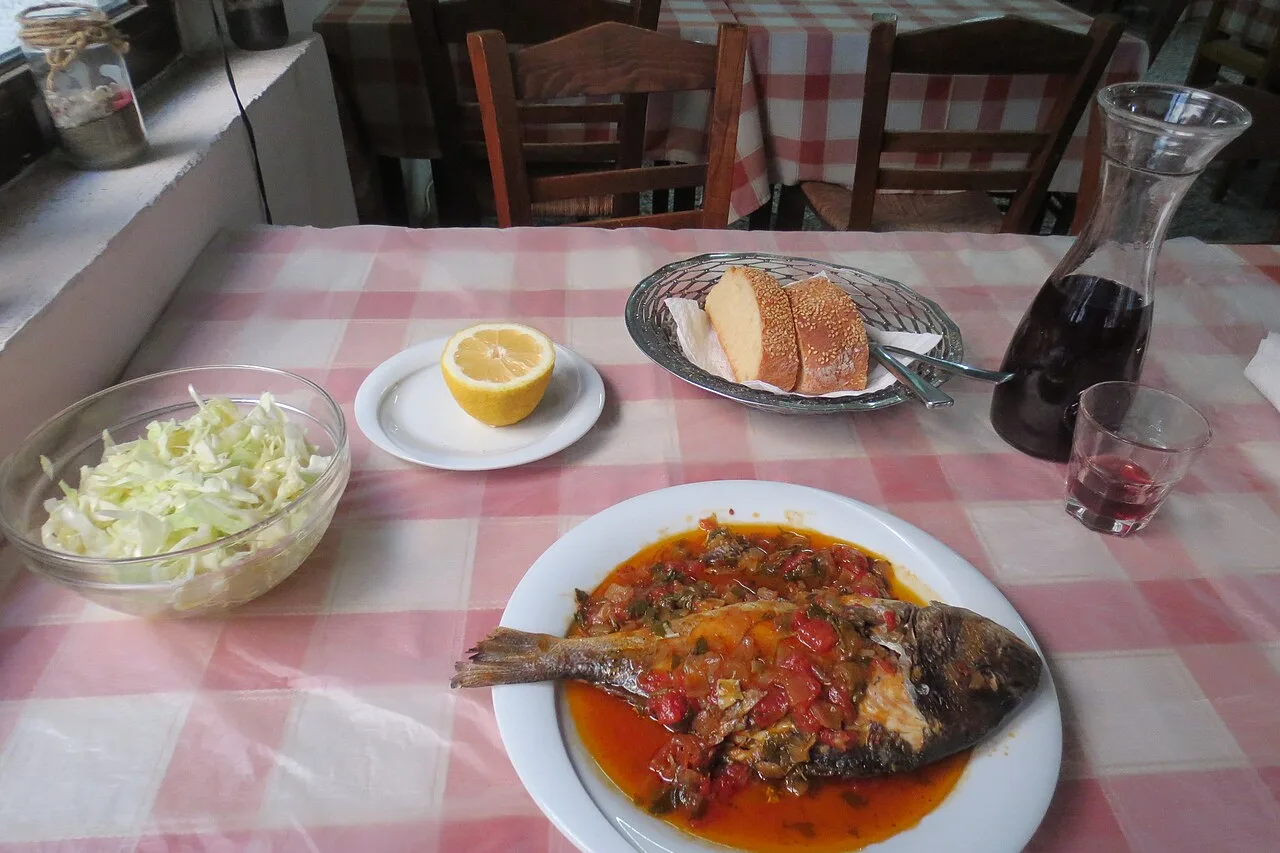 Une table de taverne grecque à nappe à carreaux : poisson à la sauce tomate, salade, pain et carafe de vin rouge