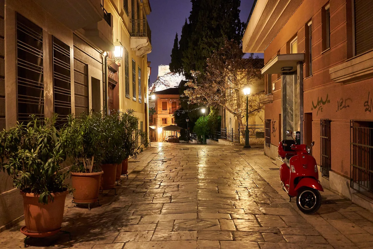 An empty paved lane in the Plaka district of Athens, lit in the evening, a red scooter on the right