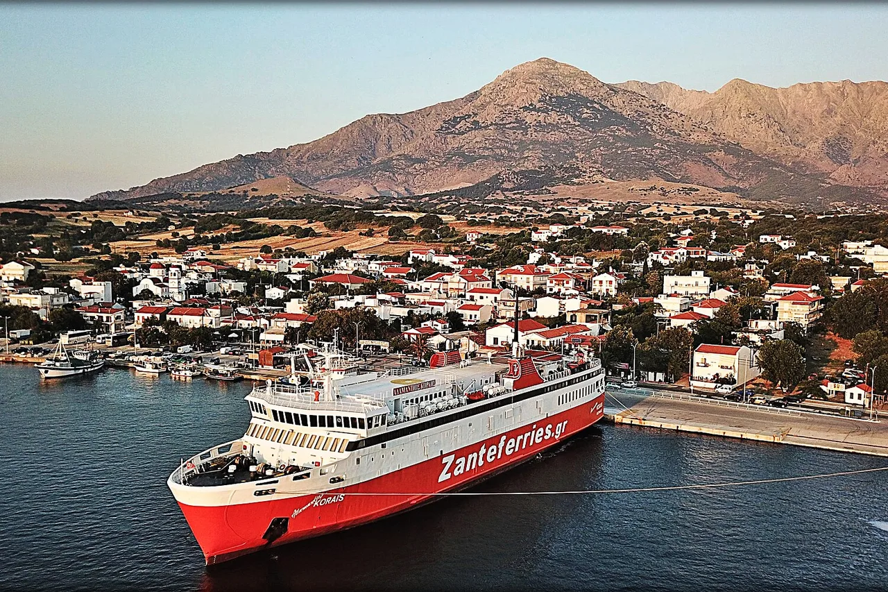 Un car-ferry rouge et blanc portant l’inscription Zanteferries est amarré dans un port devant une petite ville aux maisons blanches, avec une montagne nue derrière