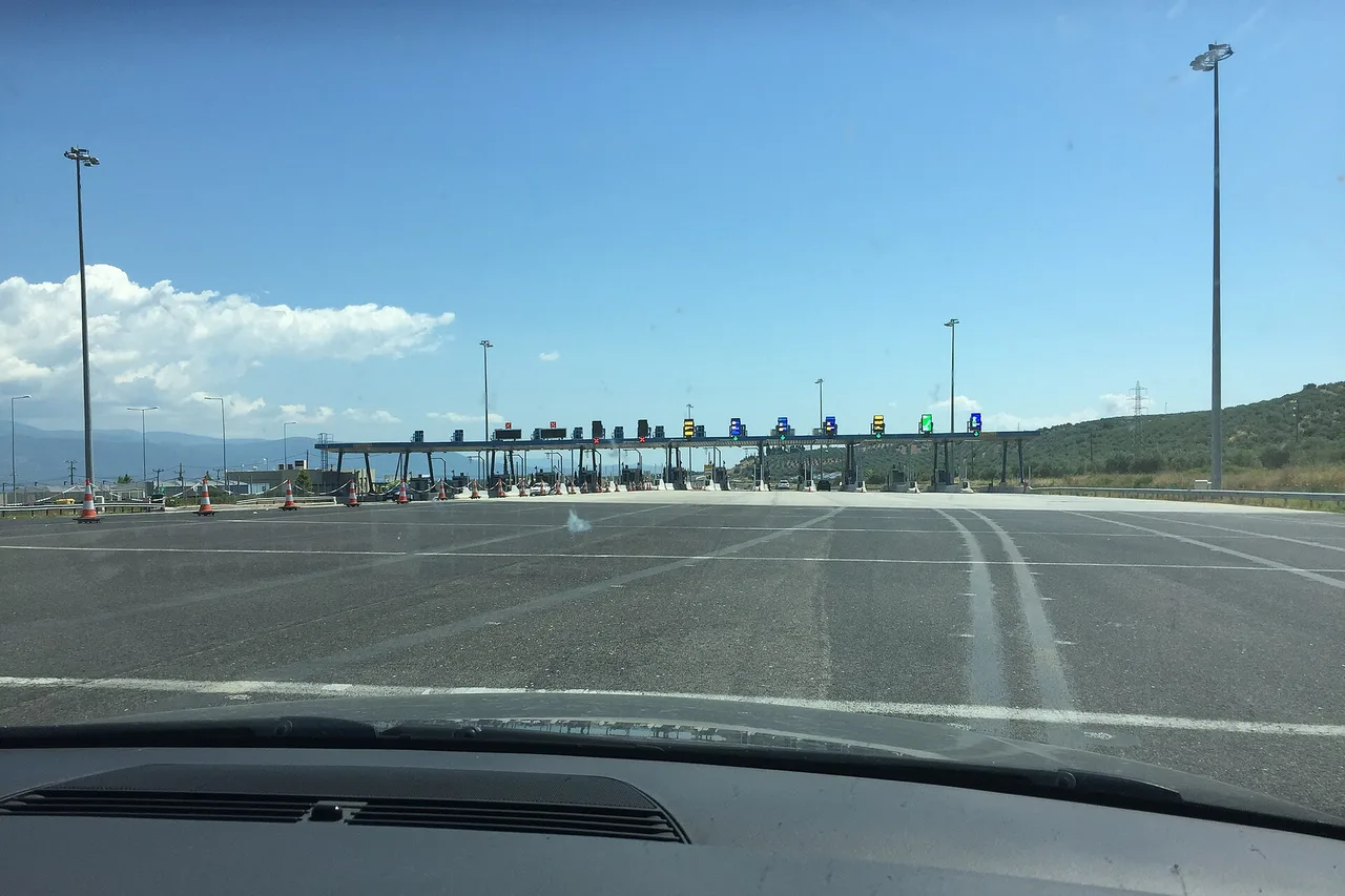 Vue à travers le pare-brise d’une voiture : une large autoroute mène à un poste de péage de nombreuses voies sous un long auvent