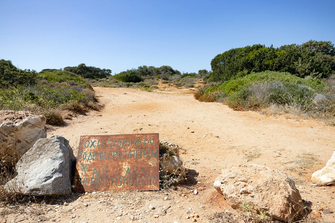 Un chemin de sable et de cailloux passe entre des buissons et des blocs de rocher ; près d’un rocher se dresse un panneau rouillé portant du texte en grec