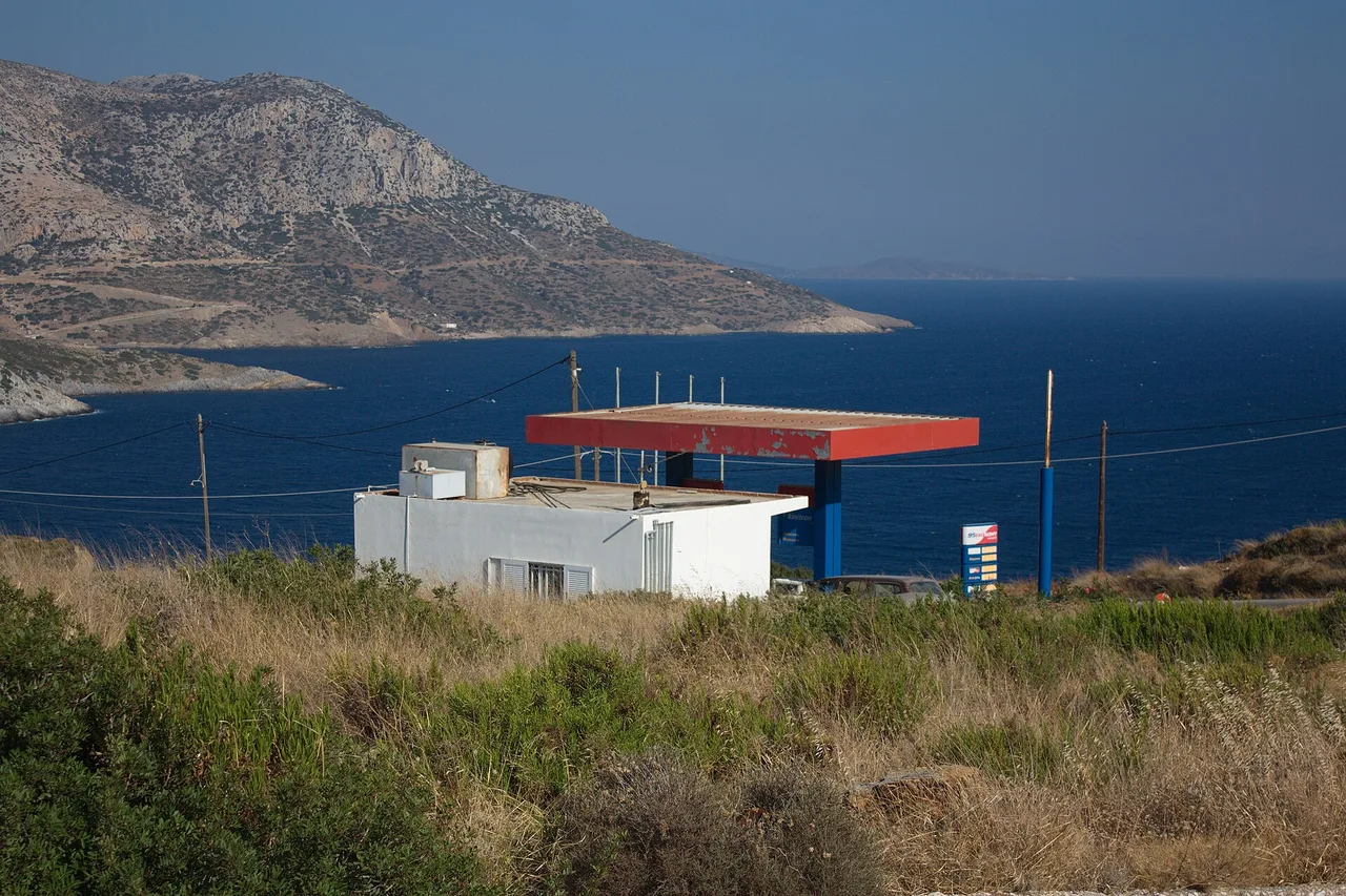 Une petite station-service au toit rouge se dresse seule sur un versant sec au-dessus d’une mer bleu foncé, avec un cap rocheux derrière