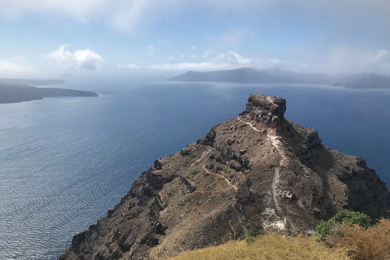 Lo sperone roccioso di Skaros vicino a Imerovigli, visto dall’alto, si protende nel mare della caldera, con isole sotto le nuvole in lontananza