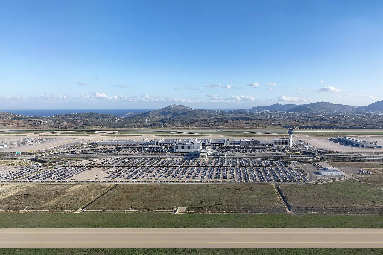 Luftbild des Flughafens Athen Eleftherios Venizelos mit Terminal, Kontrollturm und einem großen Parkplatz, dahinter Hügel und das Meer