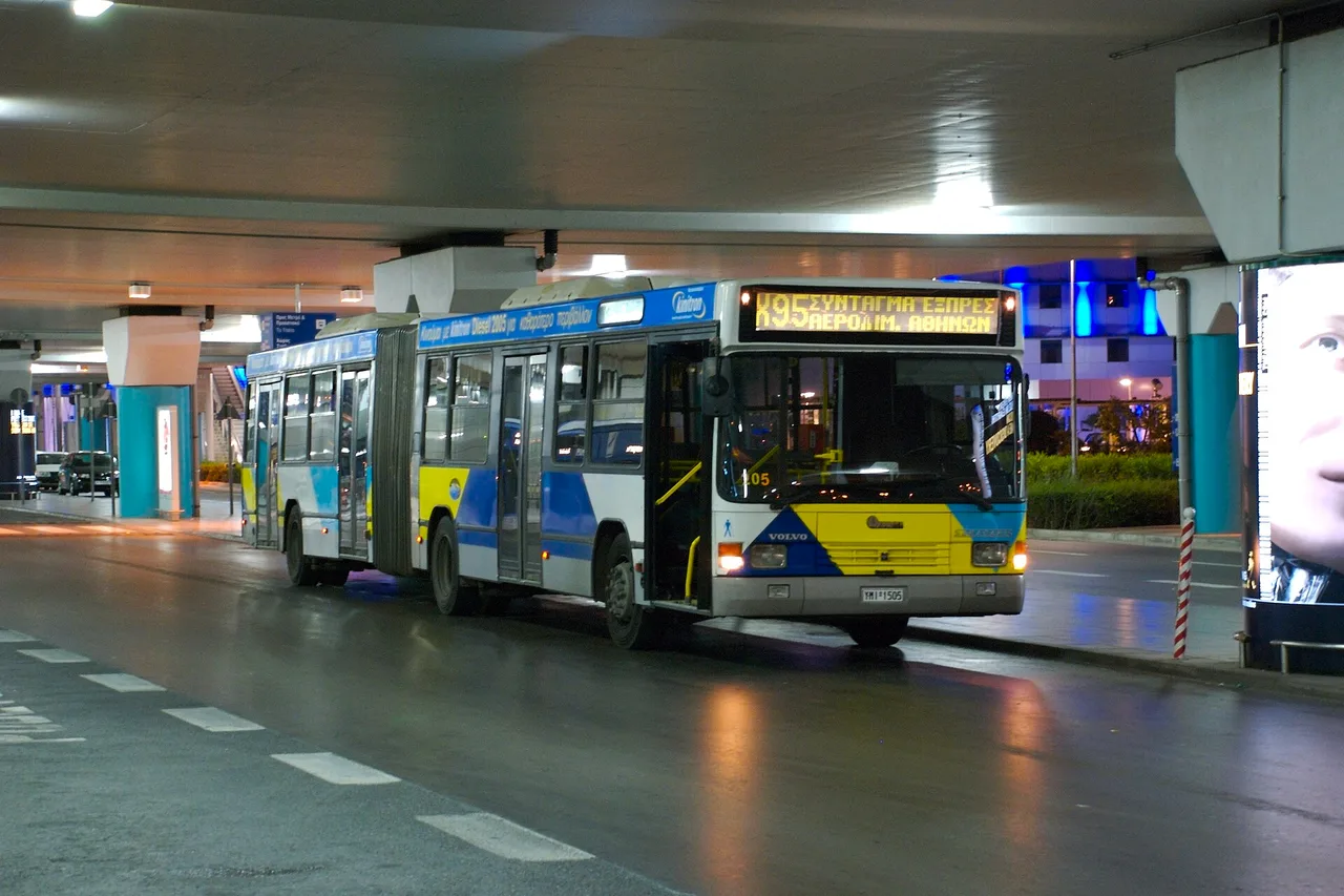 Gelenkbus der Linie X95 in den Farben der Athener Verkehrsbetriebe an der Haltestelle der Ankunftsebene des Flughafens, nachts beleuchtet