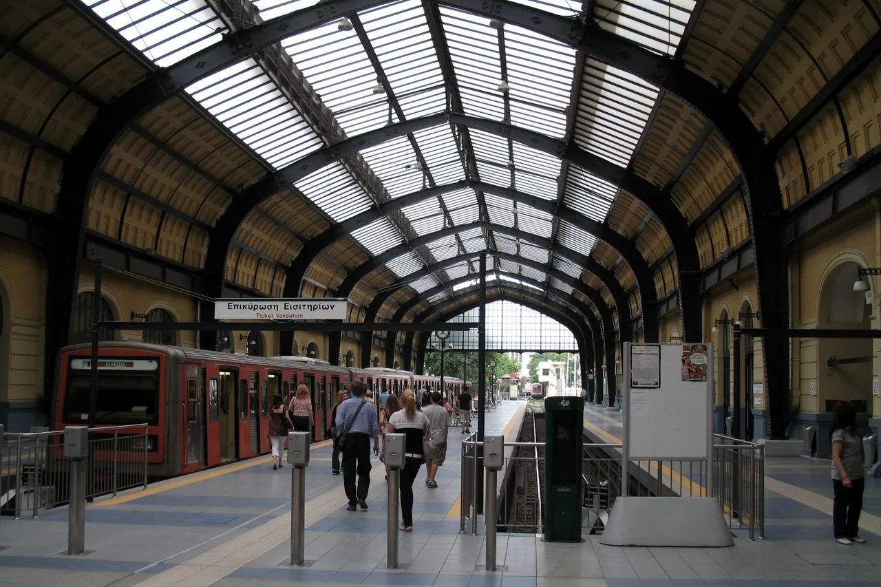 Historische Bahnhofshalle der Station Piräus mit Glasdach, Fahrgästen und einem roten Metrozug am Bahnsteig