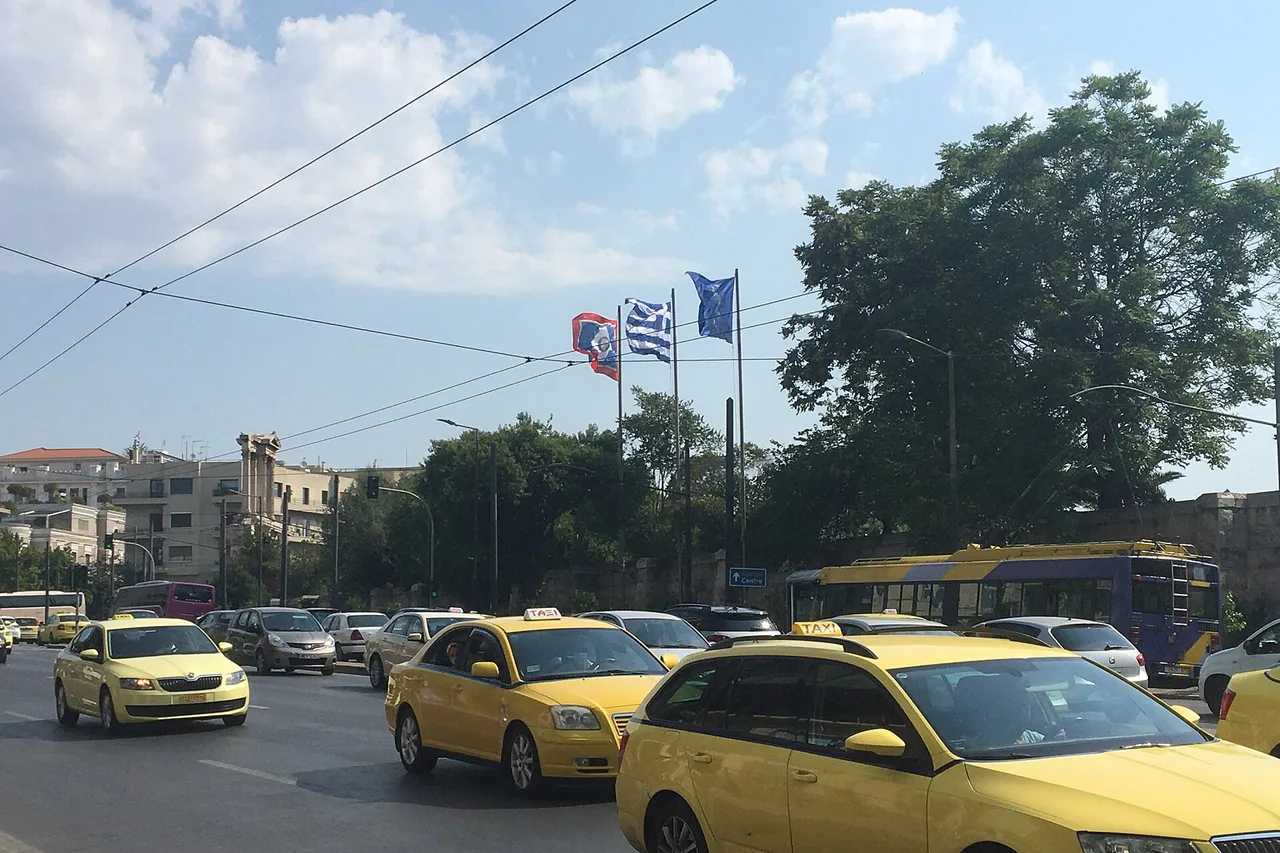 Mehrere gelbe Taxis im Verkehr einer Athener Straße unter einer griechischen und einer blauen Flagge