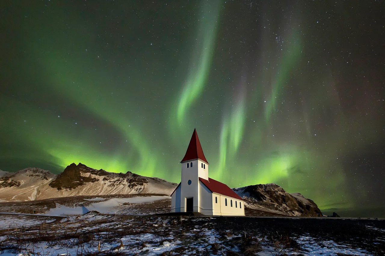 Chiesa bianca dal tetto rosso sotto un’aurora verde, paesaggio islandese innevato