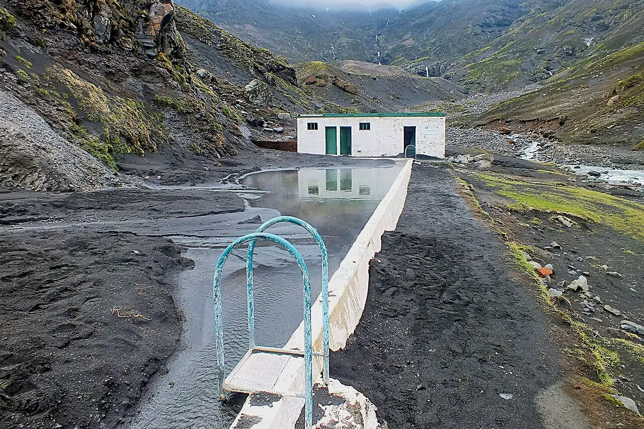 Das Becken von Seljavallalaug an der Bergwand, grünes Wasser und weiße Umkleidehütte