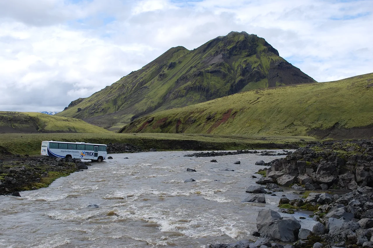 Geländebus durchquert einen isländischen Fluss unterhalb eines grünen Bergs