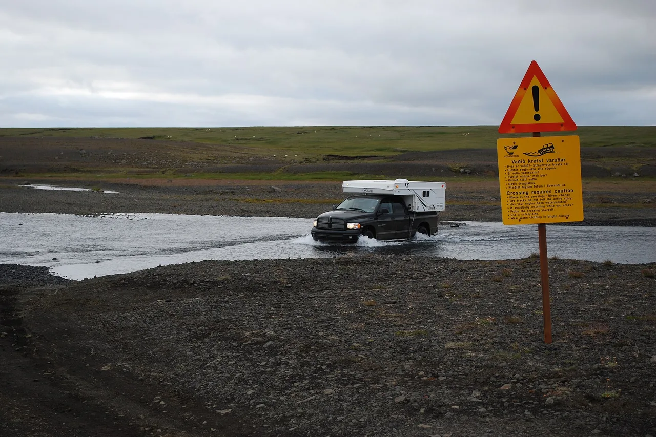 Ein schwarzer Pick-up mit Wohnkabine durchquert einen flachen Fluss, rechts davon ein gelbes Warnschild mit der Aufschrift „Crossing requires caution“