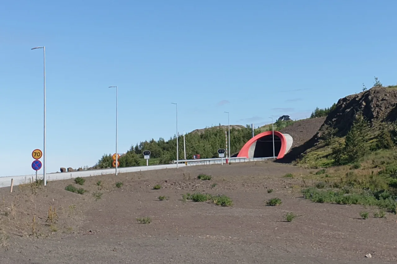 Das rot eingefasste Portal eines Straßentunnels an einem Hang neben einer Asphaltstraße mit Straßenlaternen und einem gelben Schild mit der Zahl 70