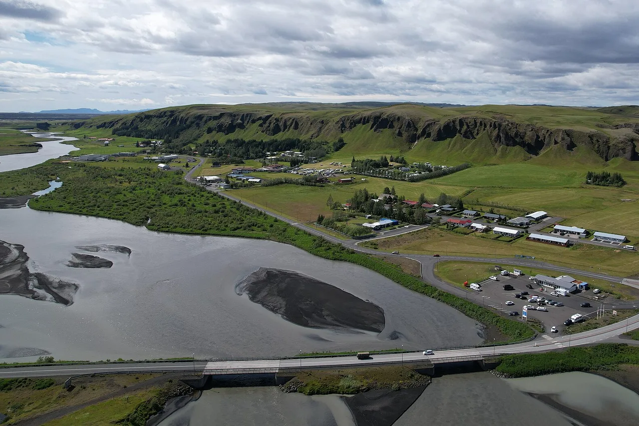 Vue aérienne de Kirkjubæjarklaustur, dans le sud de l’Islande : un petit village avec ses rues et un parking, la route circulaire franchit un pont au-dessus d’un large bras de rivière aux bancs de sable gris, derrière des falaises vertes sous les nuages.