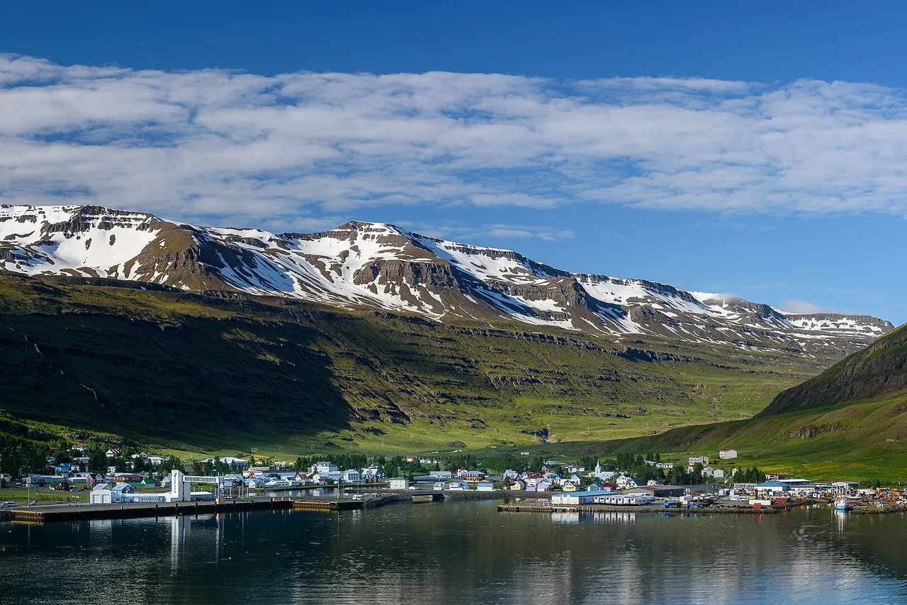 Seyðisfjörður, dans l’est de l’Islande : un village de fjord au bord de l’eau, derrière des montagnes abruptes en partie enneigées qui se reflètent dans le fjord calme.