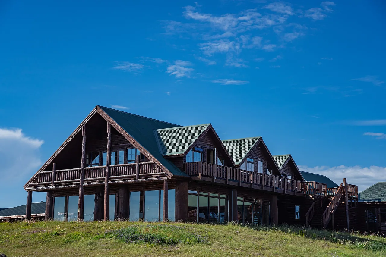 Un grand hôtel de campagne en bois, aux toits verts à pignons et aux balcons, devant une prairie et un ciel bleu.