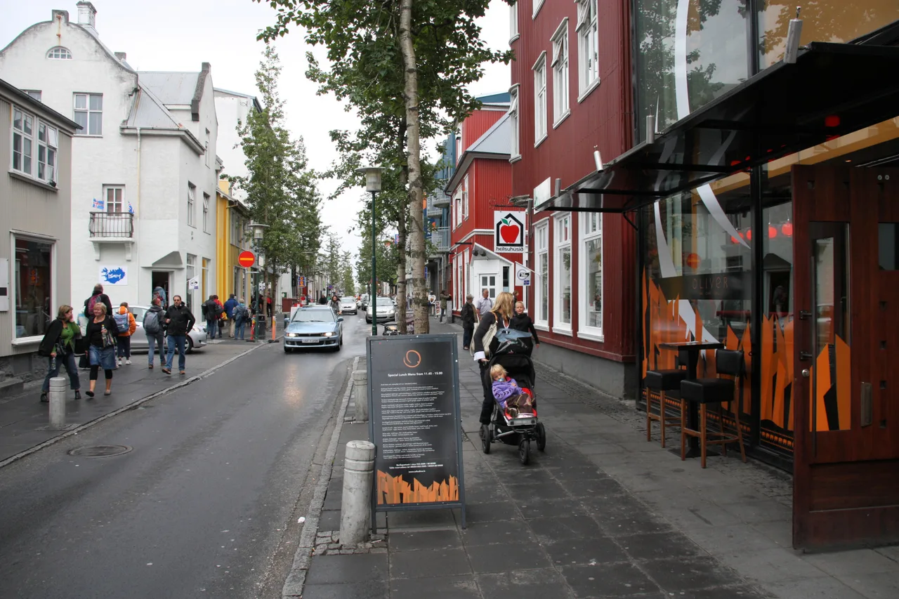 La rue commerçante Laugavegur à Reykjavík : des piétons sur les trottoirs, une voiture sur la chaussée, à gauche des maisons blanches et jaunes, à droite une maison rouge avec un café.
